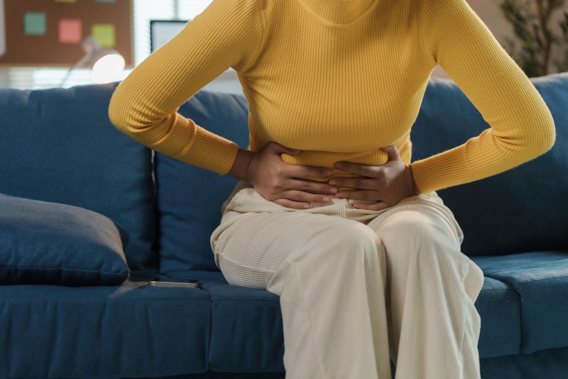 Person in a yellow sweater sits on a blue couch holding their abdomen in pain Person in a yellow sweater sits on a blue couch holding their abdomen in pain