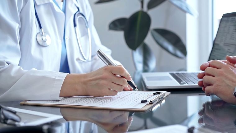 Doctor writing notes during a consultation with a patient beside a laptop Doctor writing notes during a consultation with a patient beside a laptop