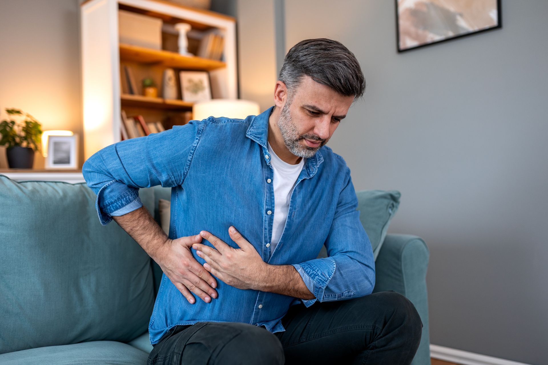 Man sitting on a couch clutching his lower side in pain in a living room