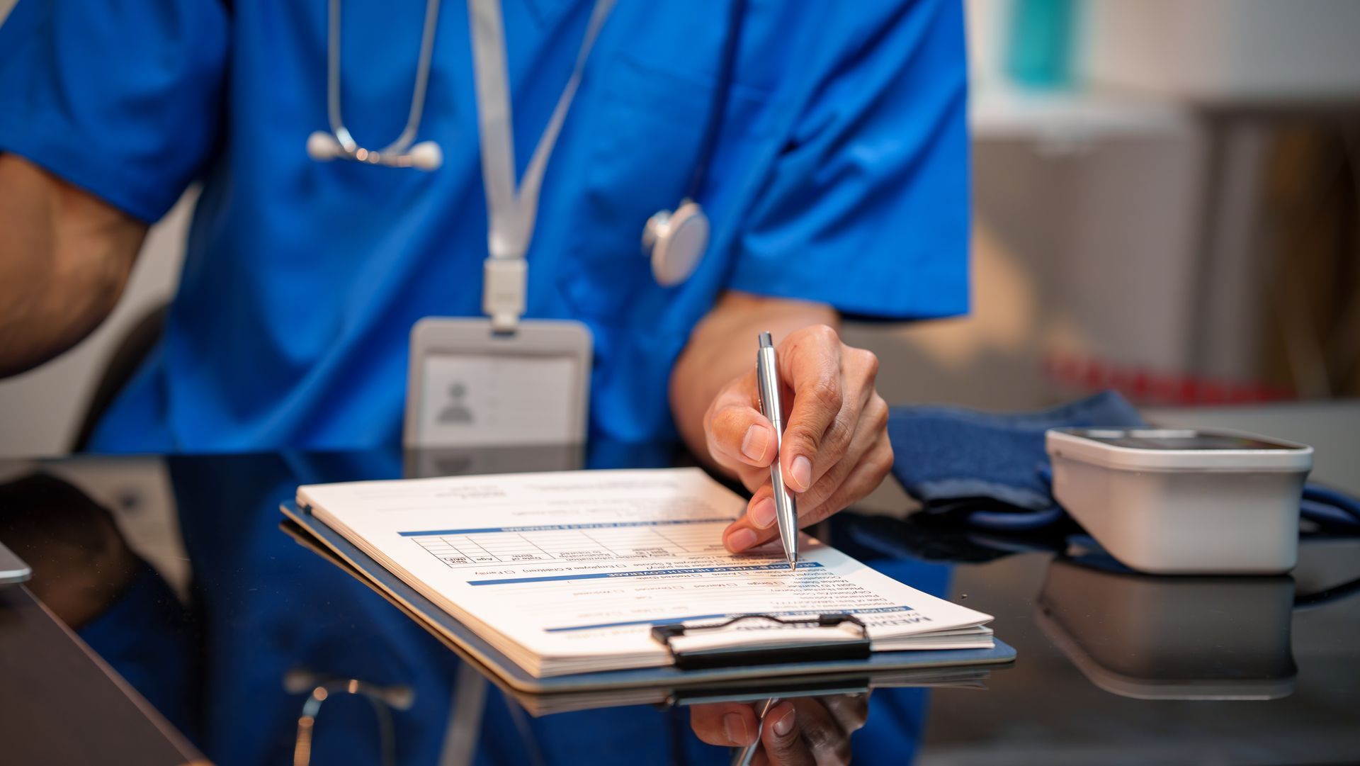 Healthcare worker in blue scrubs writing on a clipboard at a desk with a small dish nearby Healthcare worker in blue scrubs writing on a clipboard at a desk with a small dish nearby