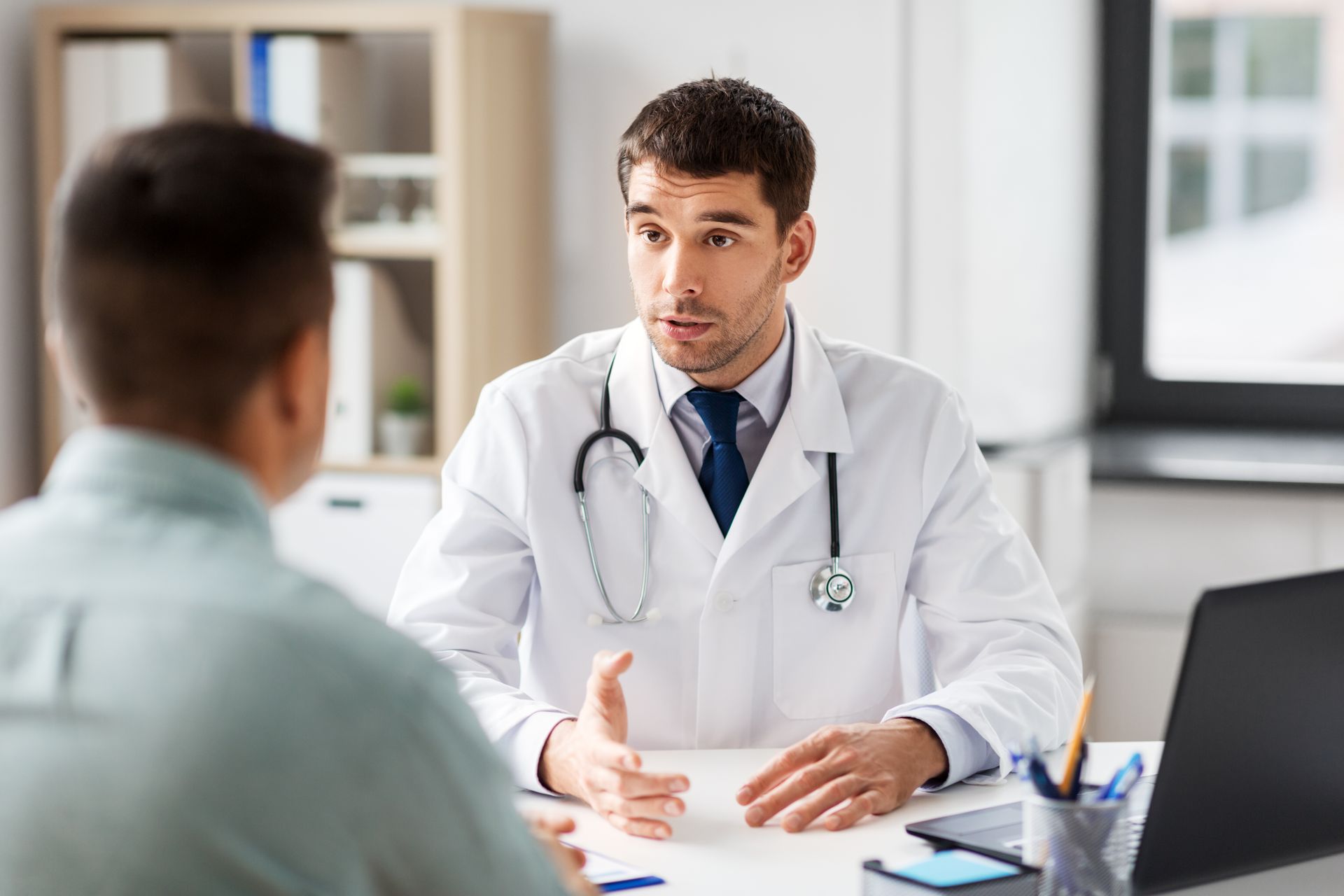 Doctor in white coat speaking with a patient in a bright office, with laptop and desk in the foreground Doctor in white coat speaking with a patient in a bright office, with laptop and desk in the foreground