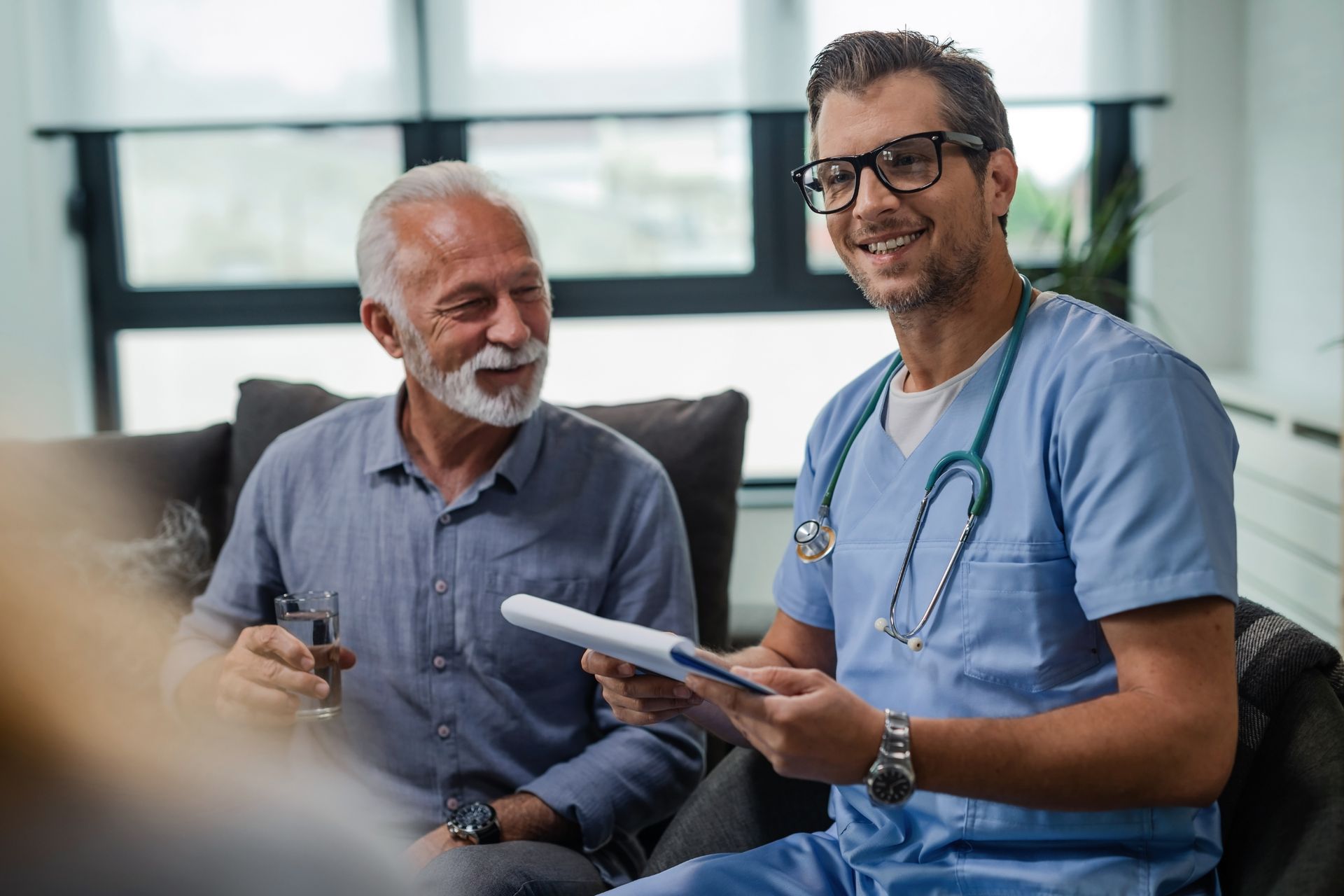 Two people in a clinic, one in blue scrubs holding a tablet and smiling while seated beside another man.