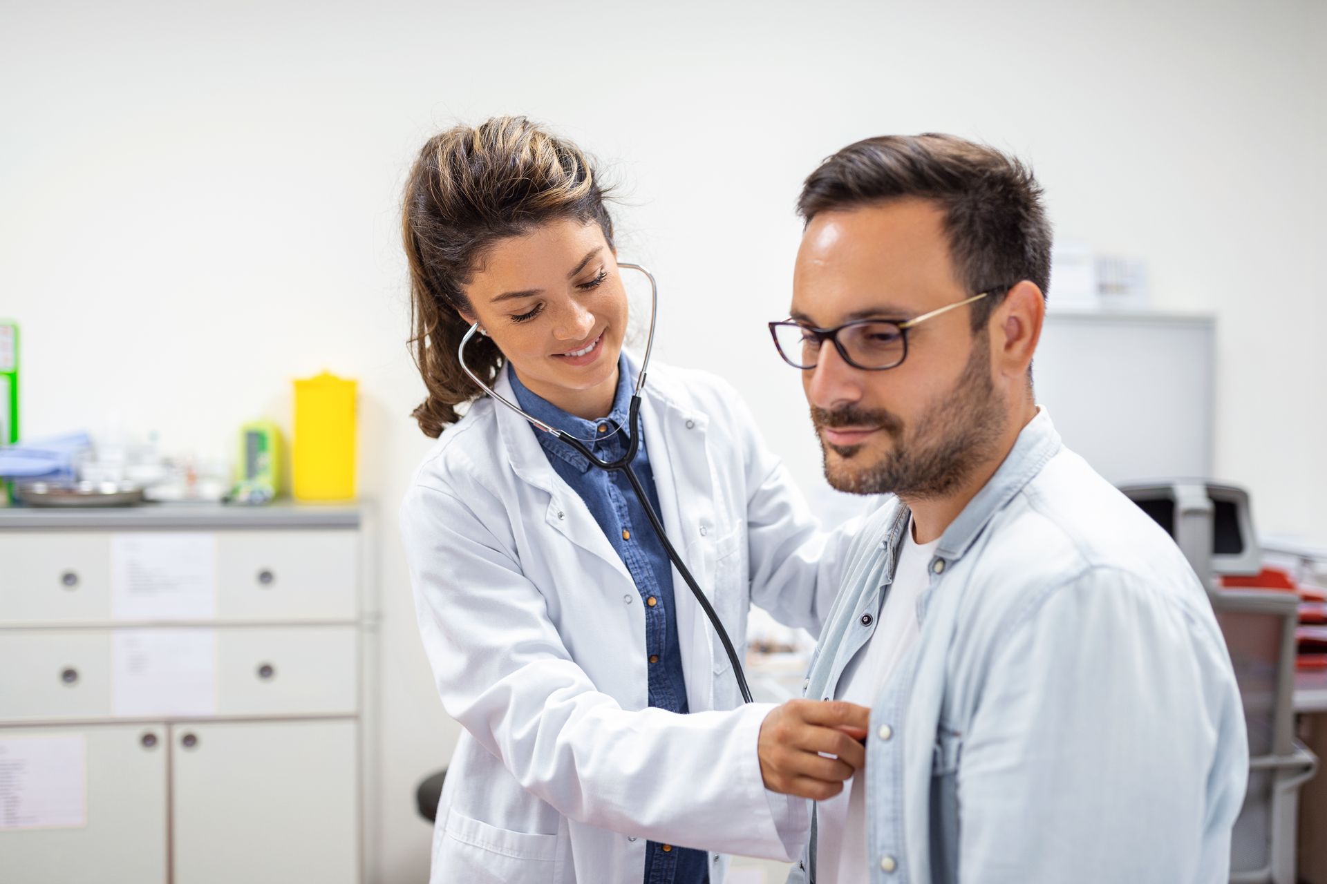 Doctor using a stethoscope to examine a seated patient in a clinic office.
