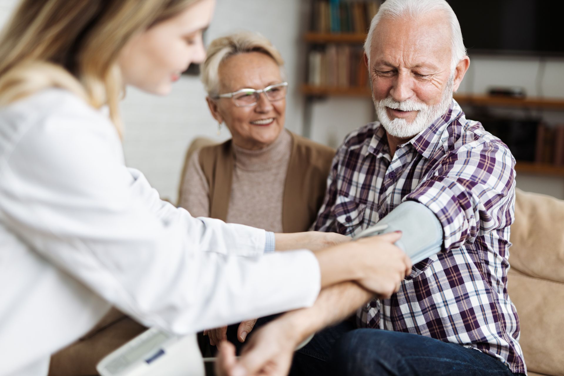 Nurse checking an elderly patient’s blood pressure while another person smiles in the background. Nurse checking an elderly patient’s blood pressure while another person smiles in the background.