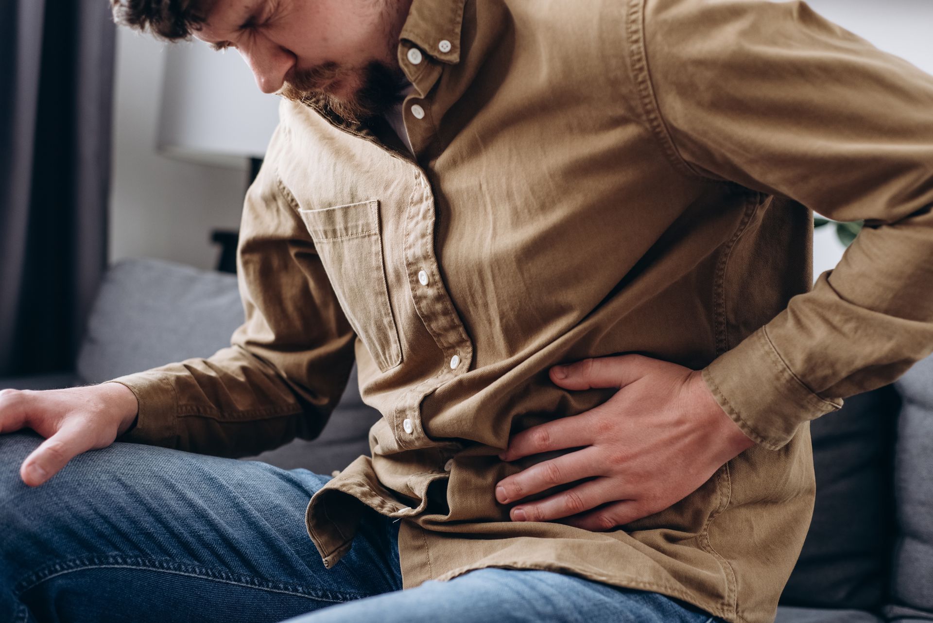 Person sitting on a couch, holding their lower back in pain, wearing a tan shirt and jeans. Person sitting on a couch, holding their lower back in pain, wearing a tan shirt and jeans.