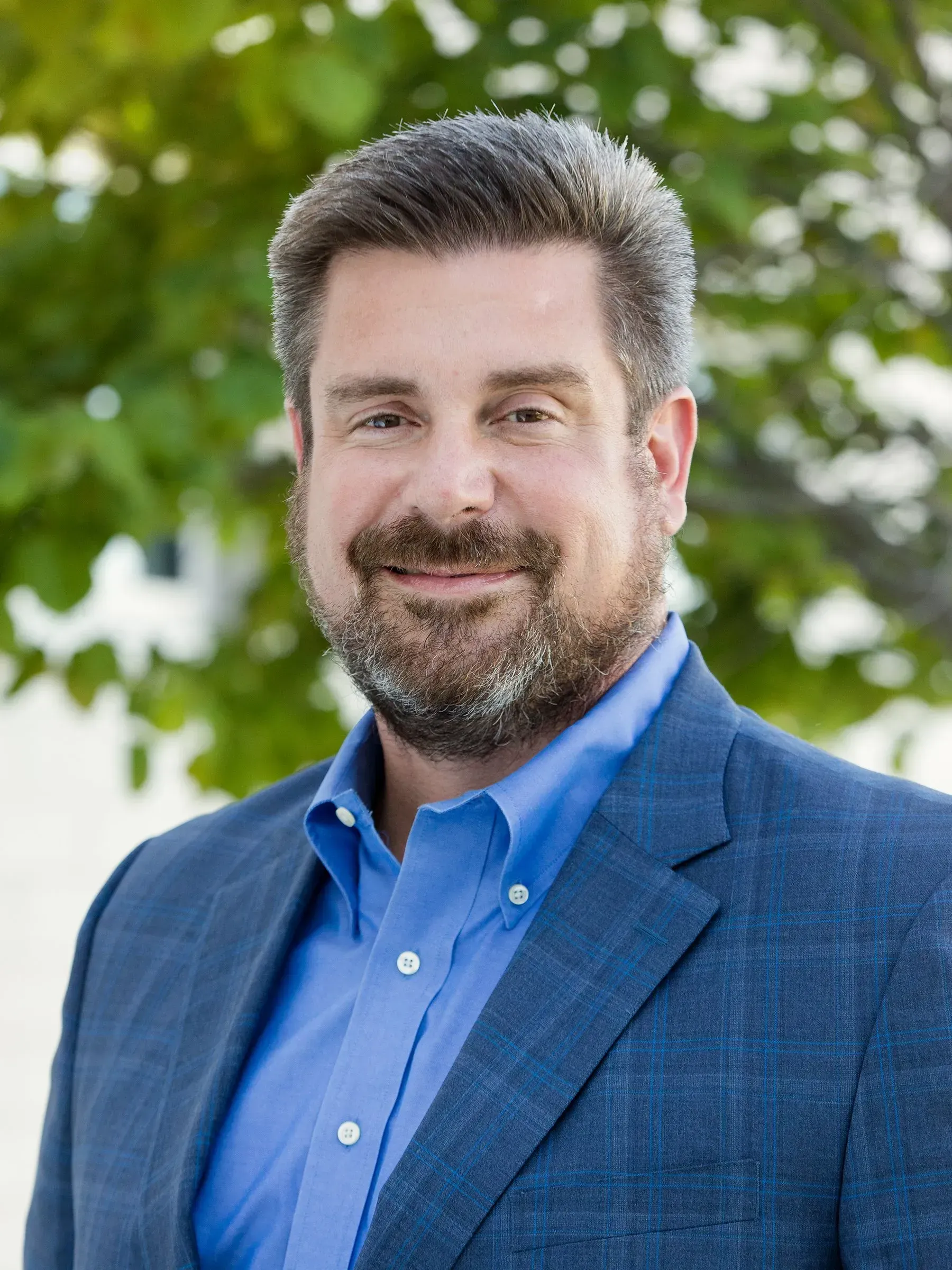 Smiling man in a blue blazer and shirt outdoors, with green trees blurred in the background Smiling man in a blue blazer and shirt outdoors, with green trees blurred in the background