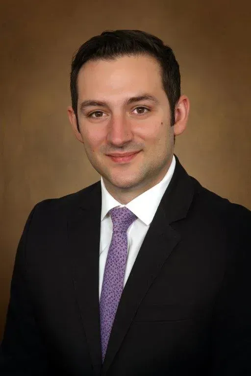 Studio headshot of a man in a black suit and purple tie against a brown backdrop Studio headshot of a man in a black suit and purple tie against a brown backdrop