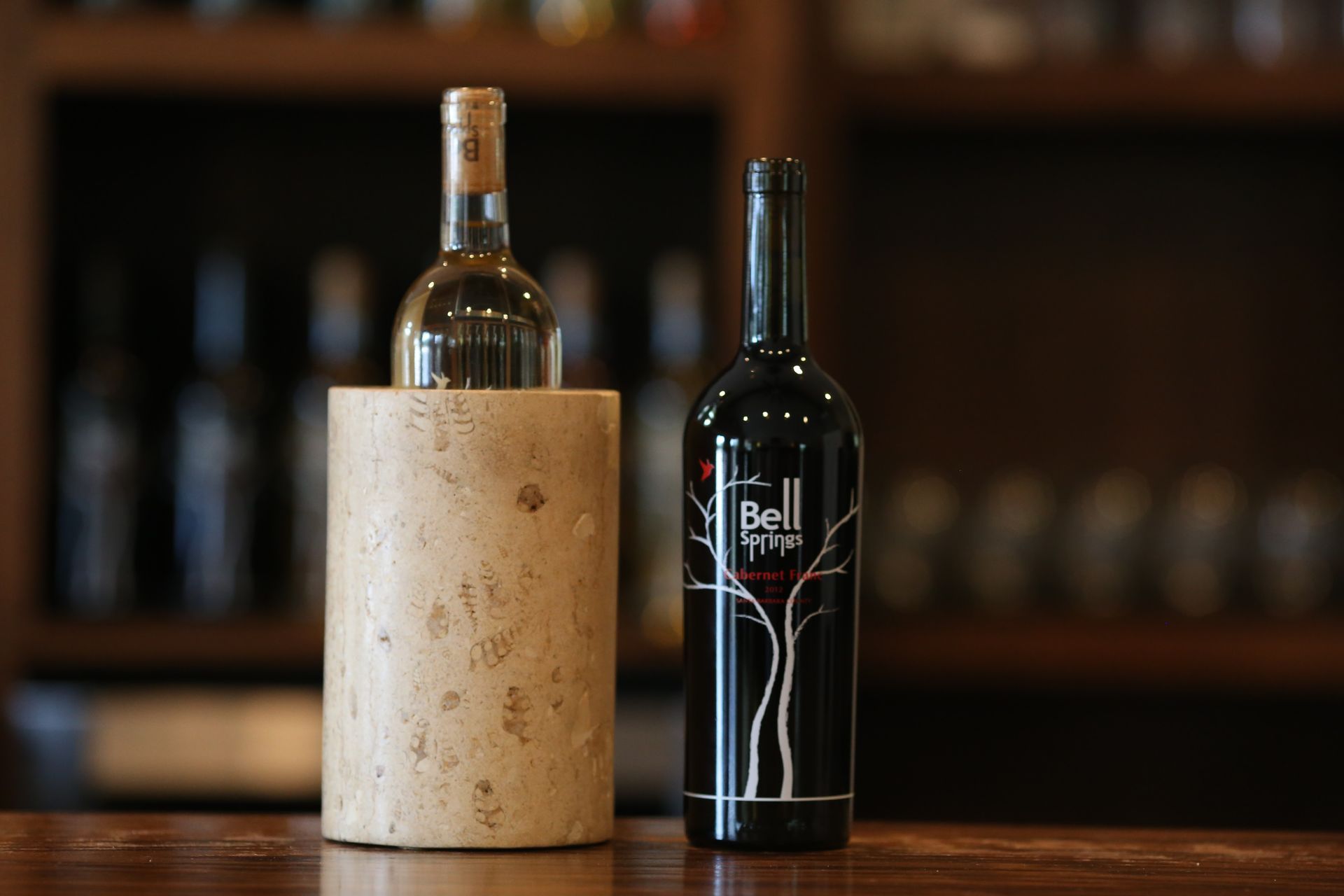 Two wine bottles: one white in a cooler, one red on a wooden bar with a blurred background.