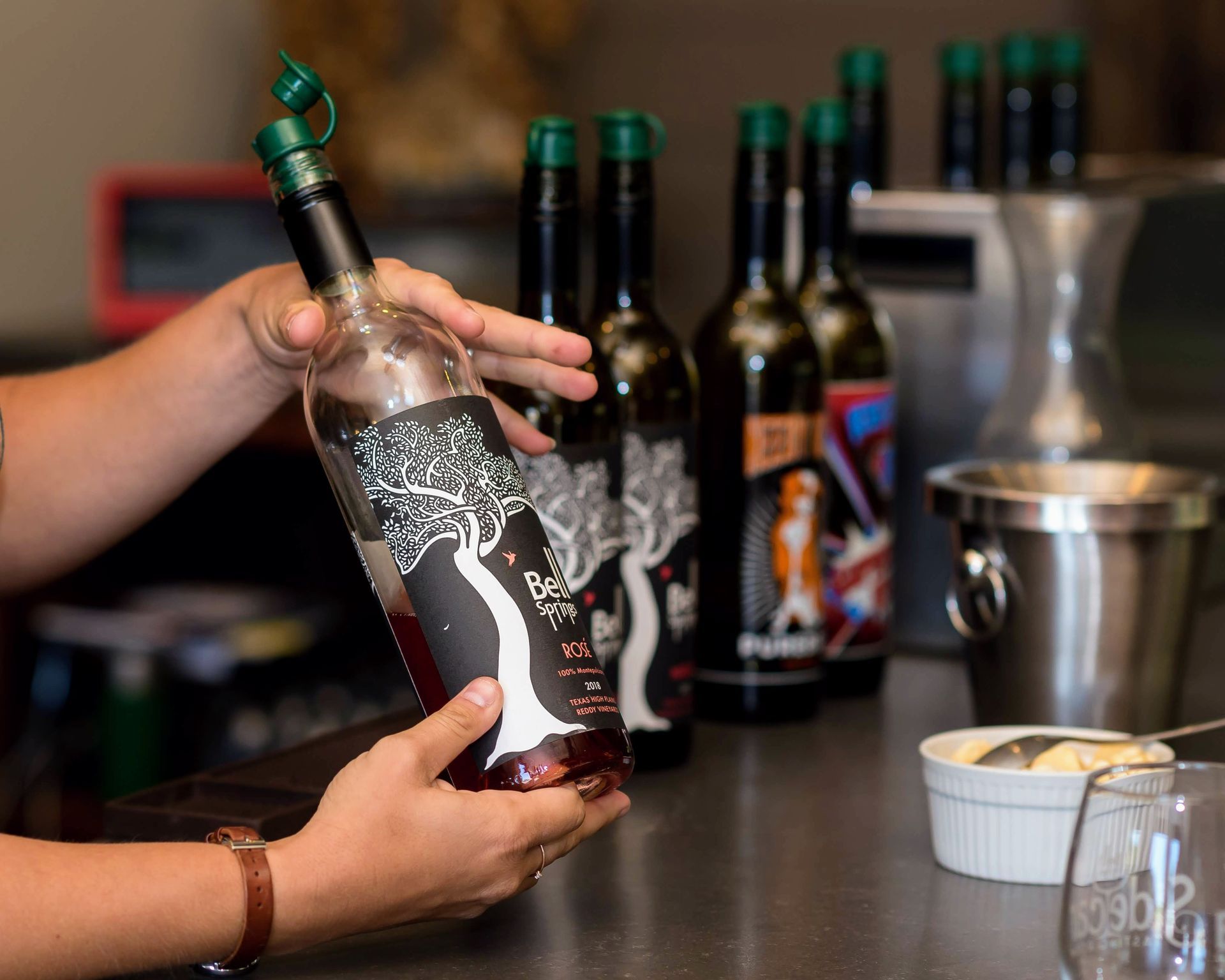 Person holding a bottle of dark liquid, other bottles and a bowl of white substance on a counter.