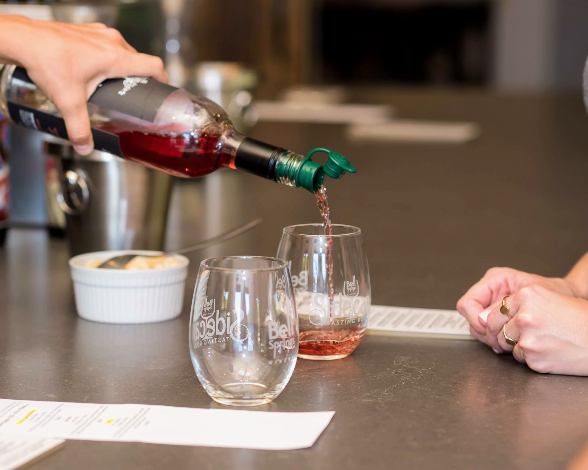 A hand pouring red wine into a glass at a tasting. Another glass and two people are also present.