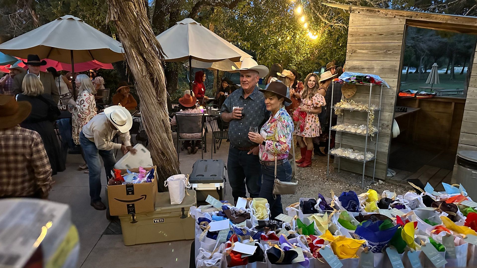 Outdoor fair: People in cowboy hats, browsing merchandise at tables. Warm lighting, evening setting.