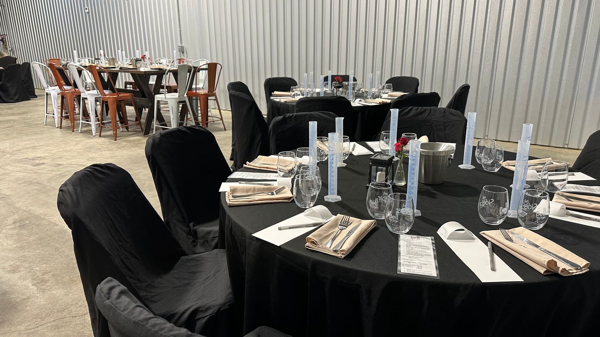 Round tables set for dining, covered in black with black chair covers. Tables are in a warehouse.