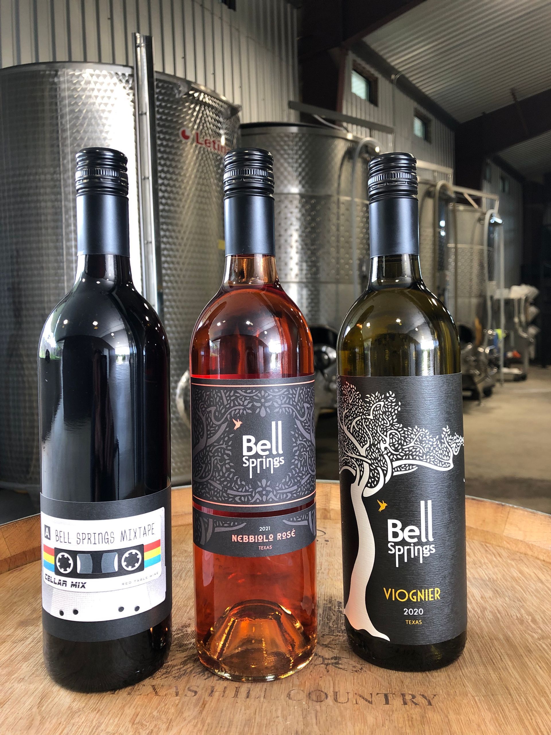 Three bottles of wine: red, rosé, and white, on a wooden surface. Stainless steel tanks are in the background.