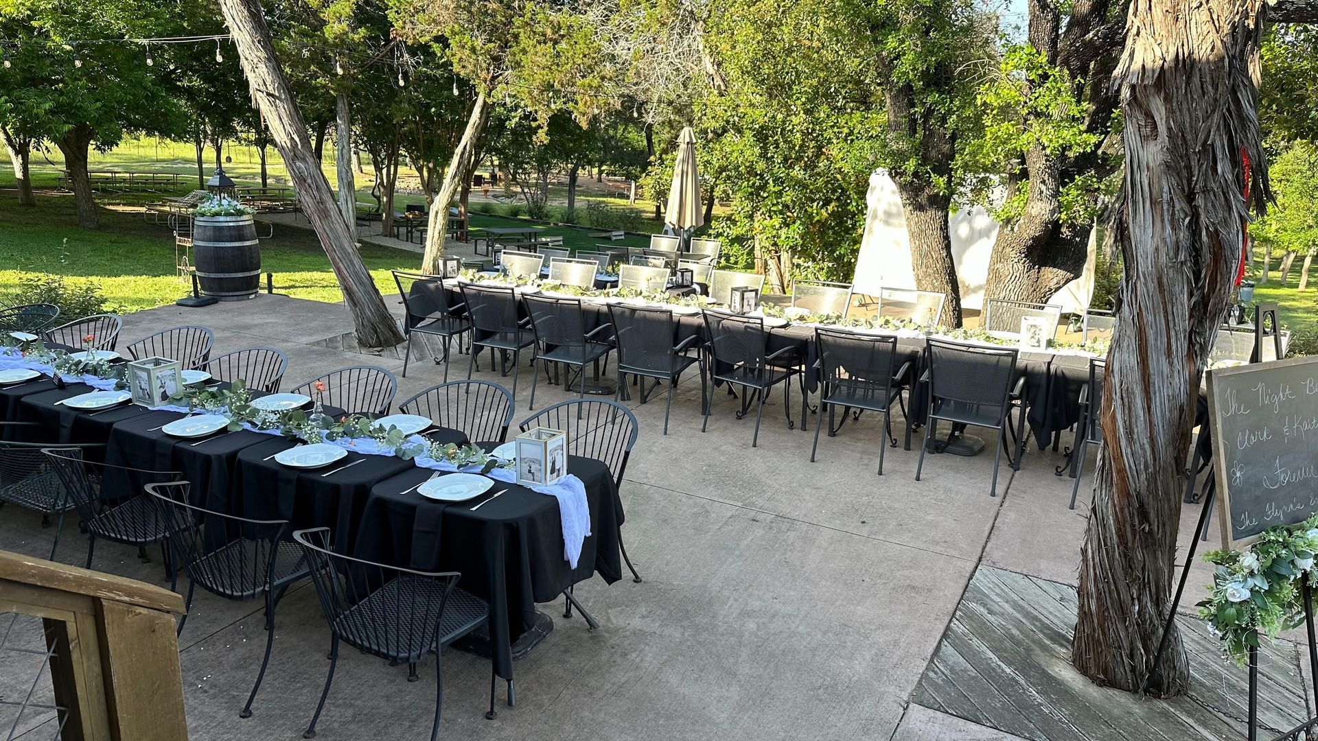Long tables set outdoors, black tablecloths, metal chairs, ready for a gathering under trees, sunny day.