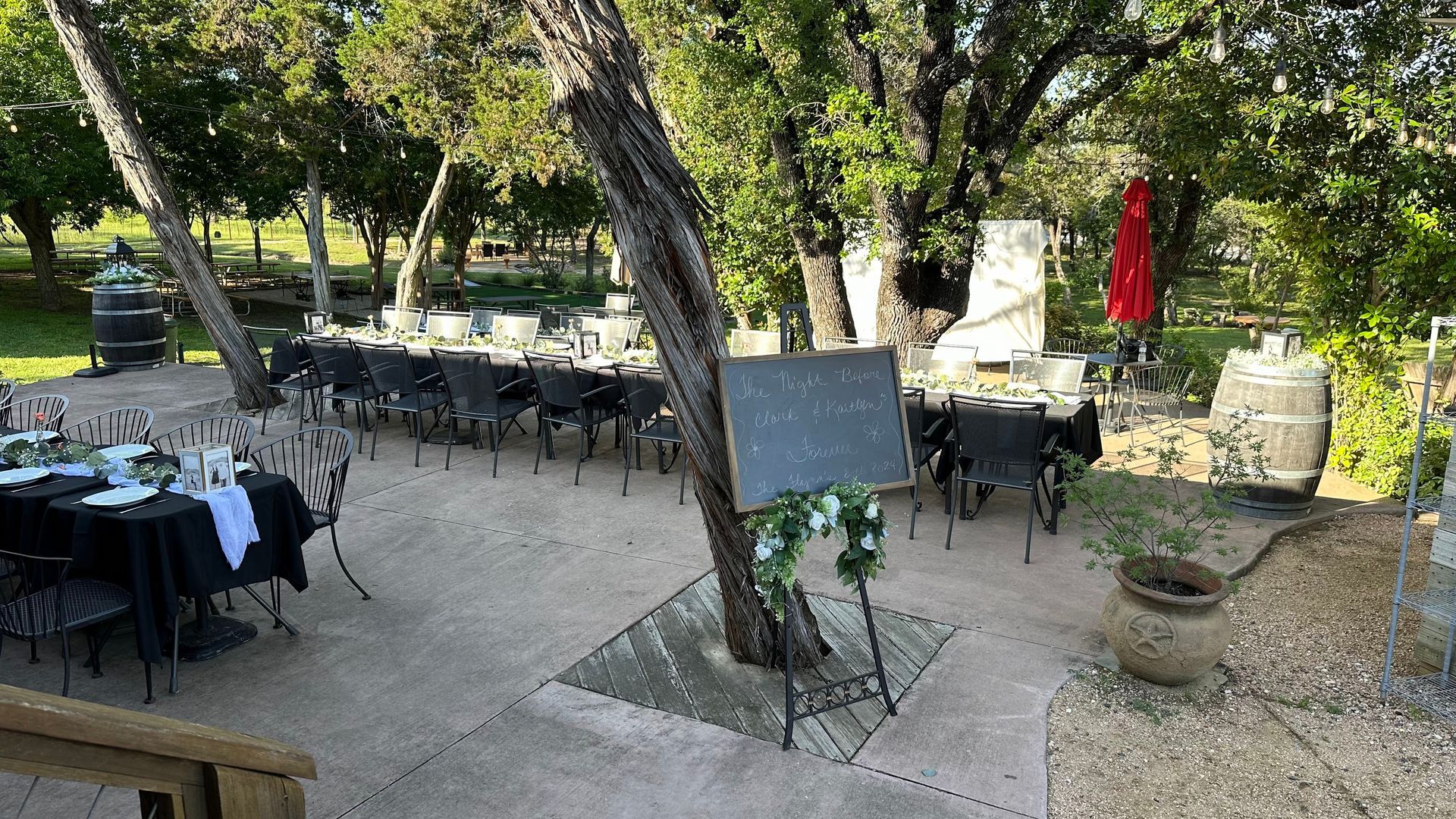 Outdoor dining tables set for a celebration under trees, with a chalkboard menu.