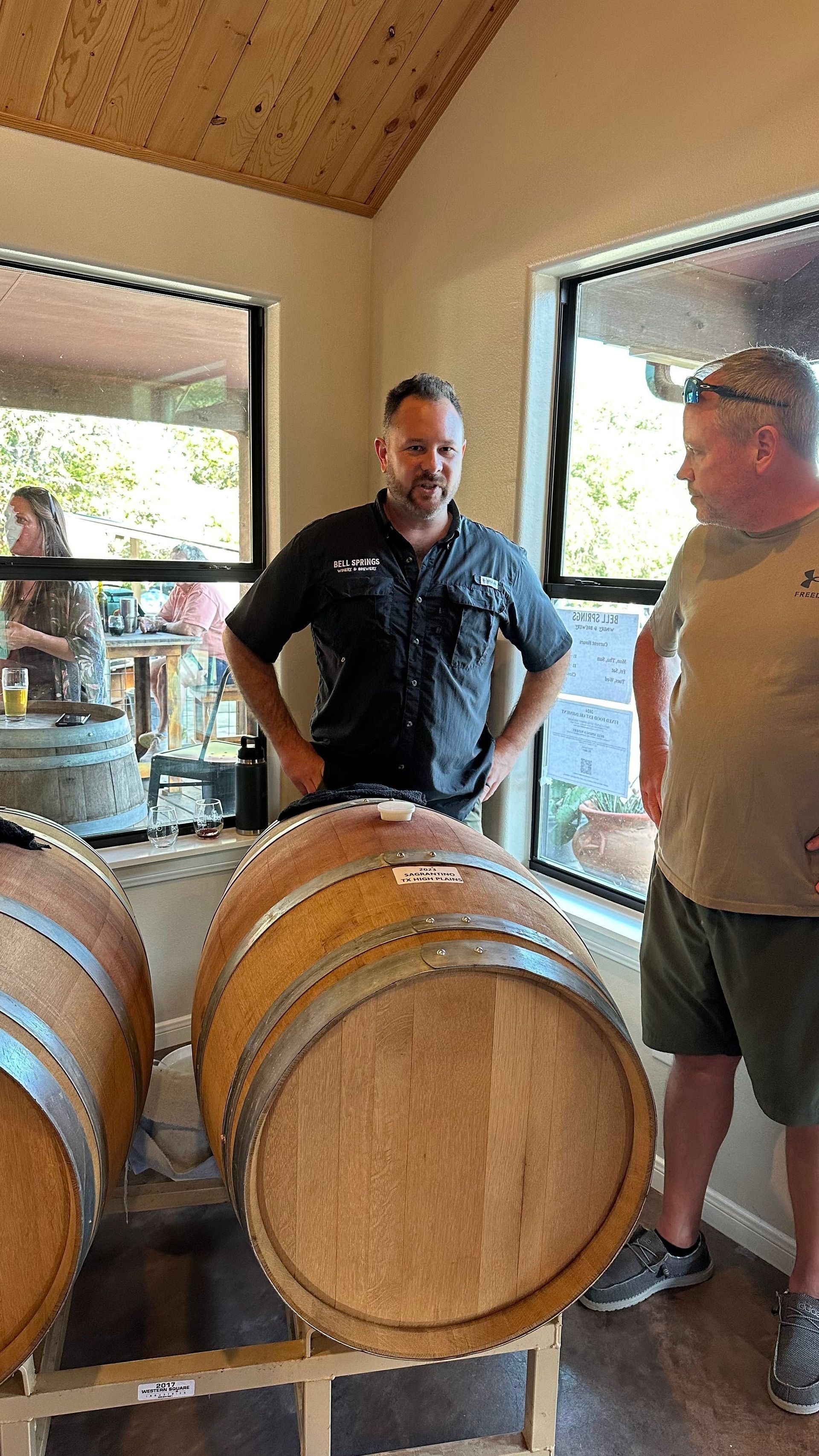 Two men standing by wine barrels in a tasting room. One is talking, another is listening.