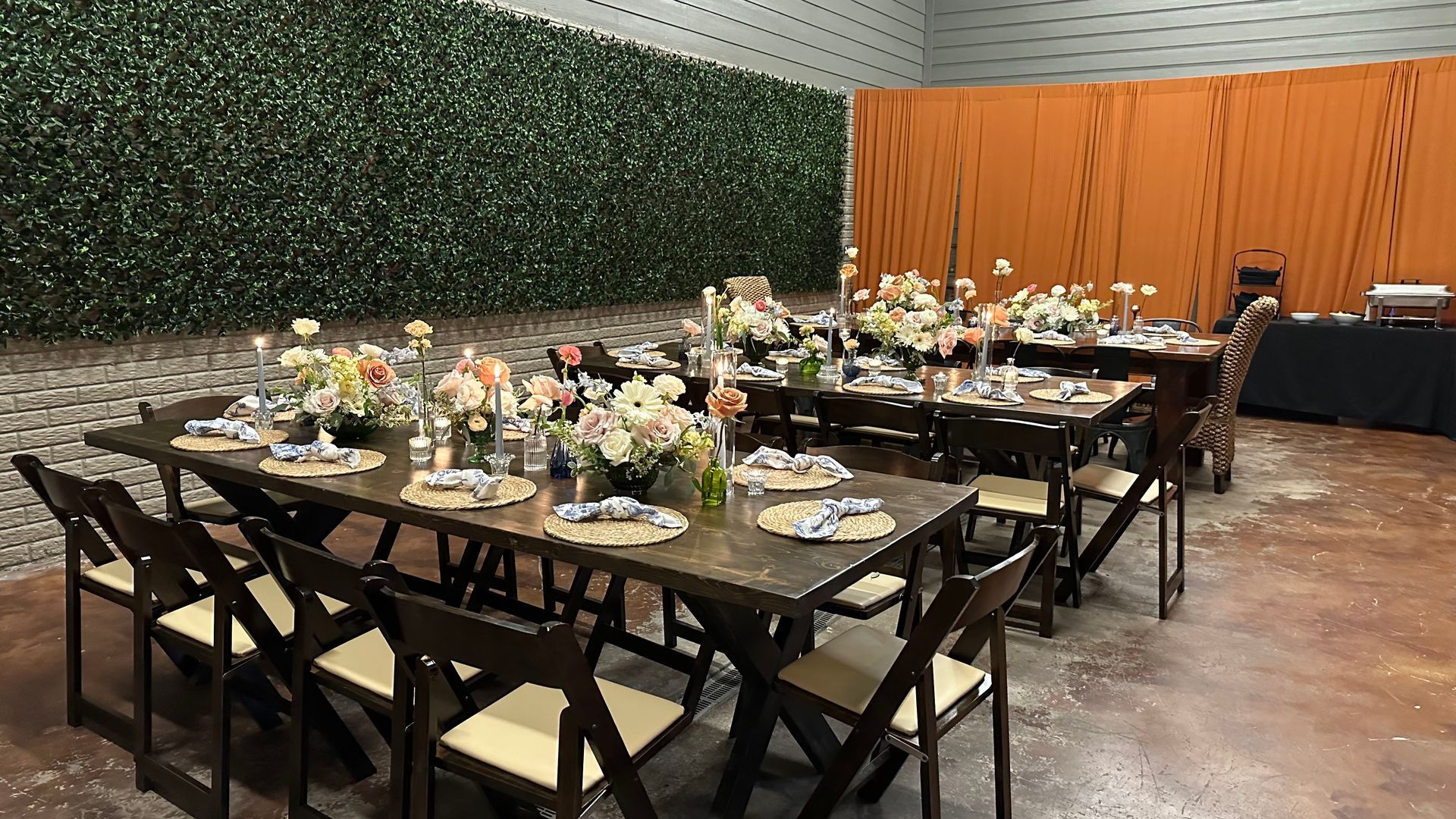 Tables set for a formal event, flowers and place settings arranged on dark wood. Dark chairs and a green wall.