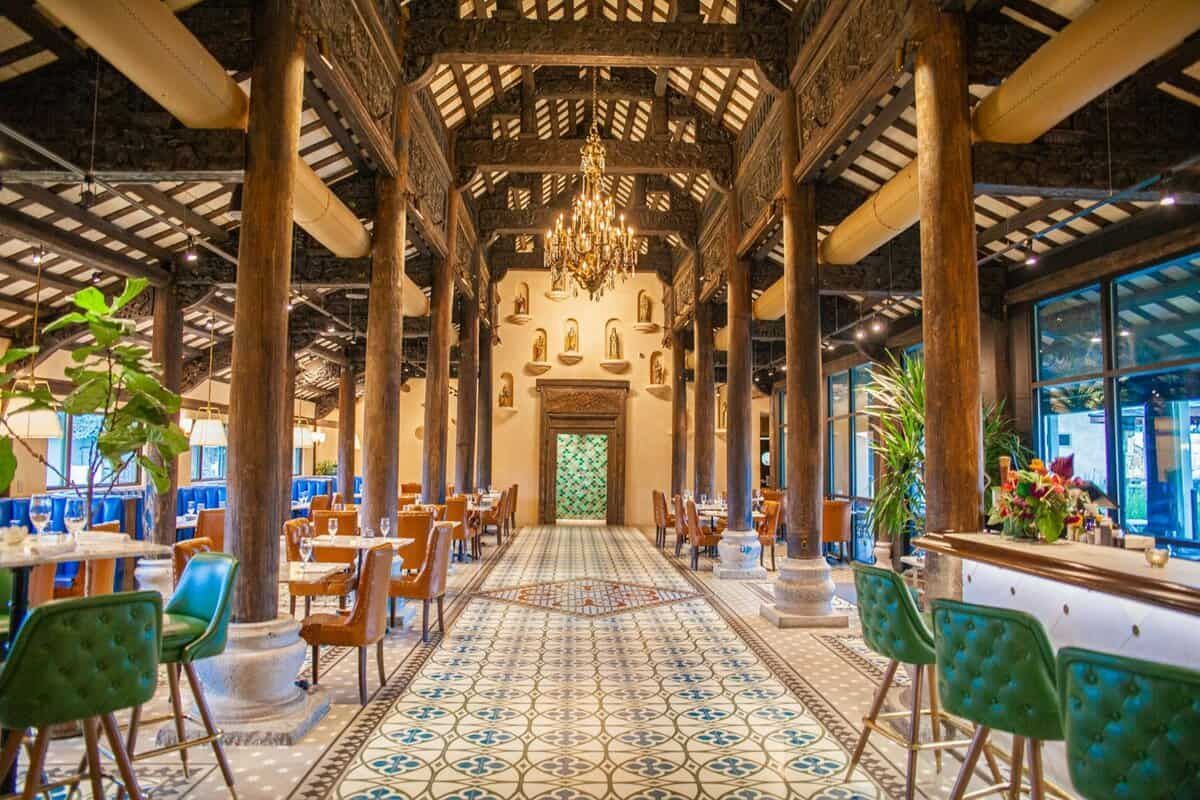 Ornate, open-air restaurant with high ceilings, wooden beams, and a patterned floor. Tables, chairs, and bar stools fill the space.