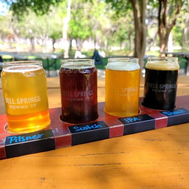 Flight of beers in a wooden holder: Pilsner, Scotch, IPA, and a dark beer, outdoors.