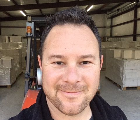 Man smiling in warehouse, forklift in background, surrounded by boxes.