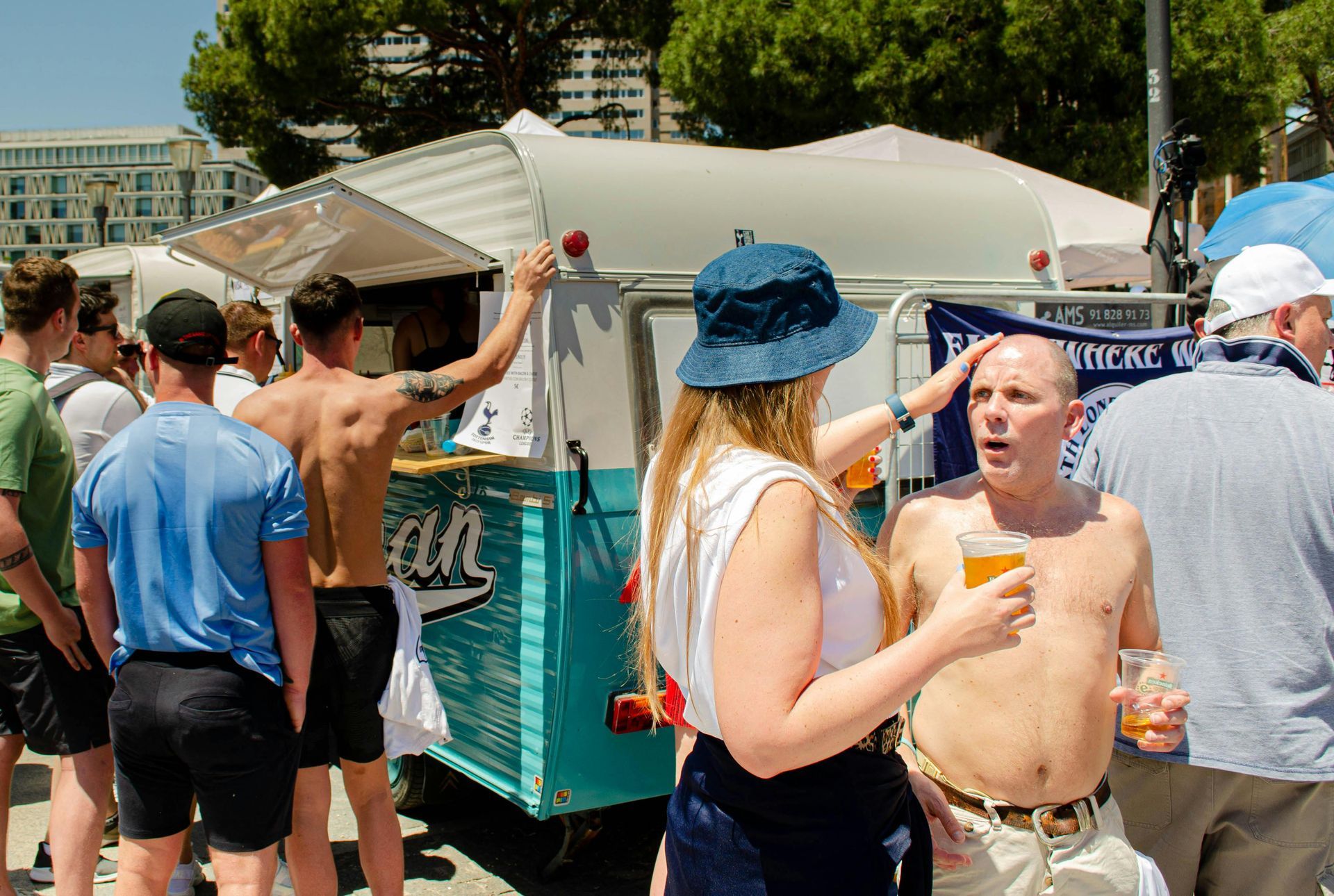 People at an outdoor bar, some shirtless, drinking beer. Woman touches a man's head, sunny day.