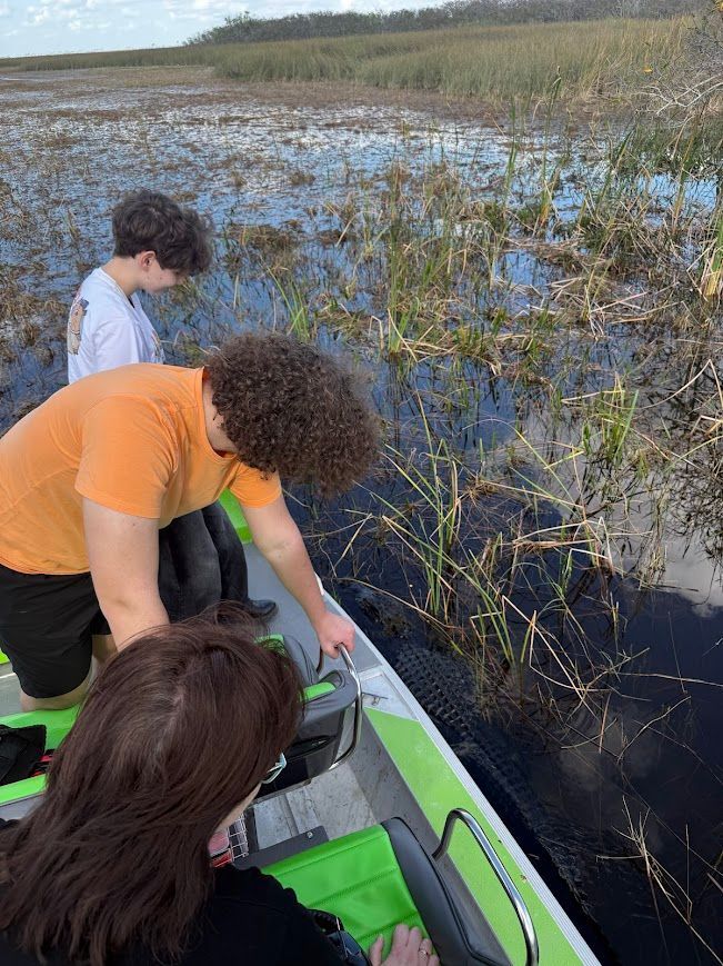 Three people in a small boat observe aquatic plants in a wetland. The person in front has long brown hair.