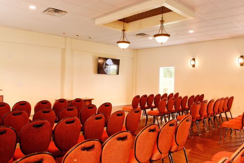 Rows of red chairs in a bright conference room with ceiling lights and a wall-mounted TV