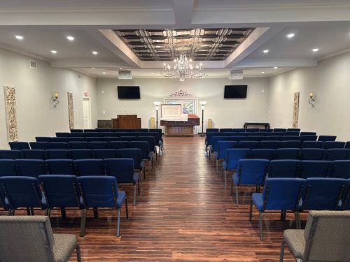Empty chapel with rows of blue chairs, a polished wood aisle, and a chandelier under a white ceiling.