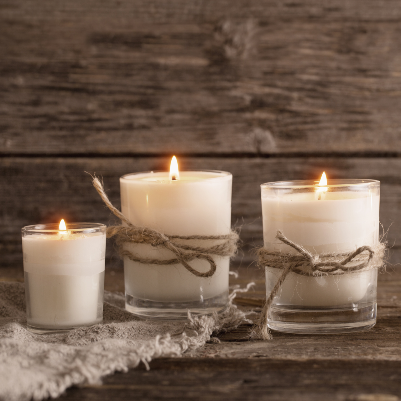 Three lit white candles in glass jars tied with twine on a rustic wooden table, with a lace cloth nearby