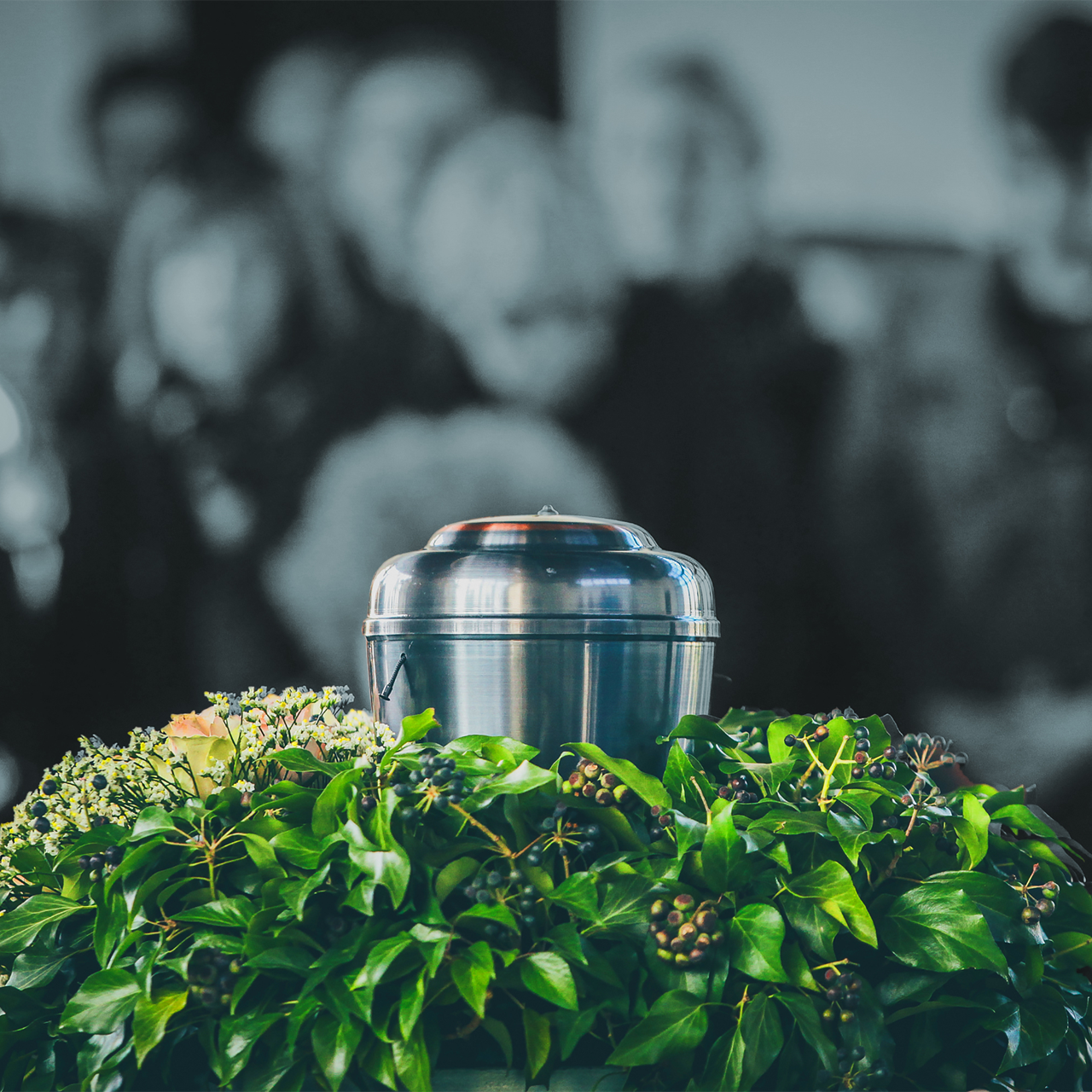 A silver urn among green flowers with a blurred crowd in the background