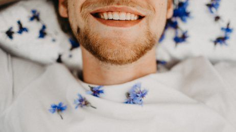 Smiling person in a white sweatshirt with blue floral details, lying on a bed or couch