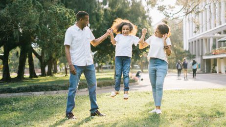 Family jumping together in a sunny park, holding hands on the grass near trees and a building.