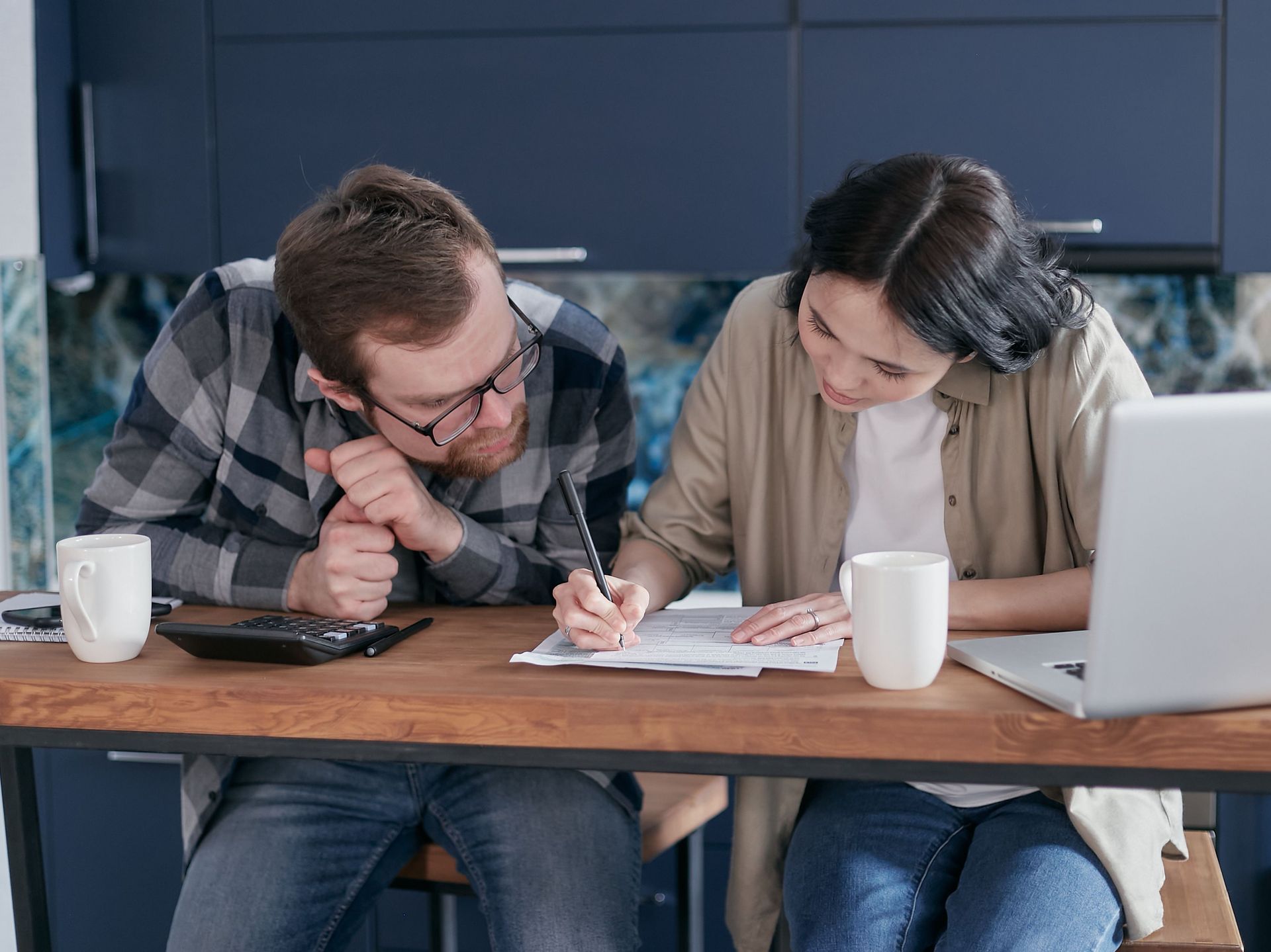 Two coworkers leaning over paperwork at a kitchen table with mugs and a laptop