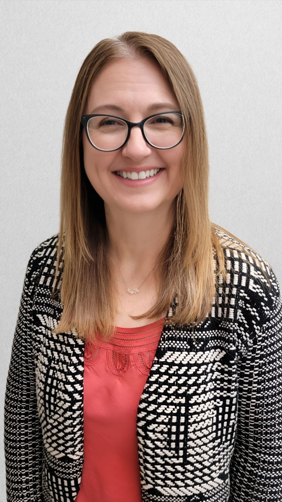 Woman with glasses, smiling, wearing a patterned jacket over a red top, set against a white wall.