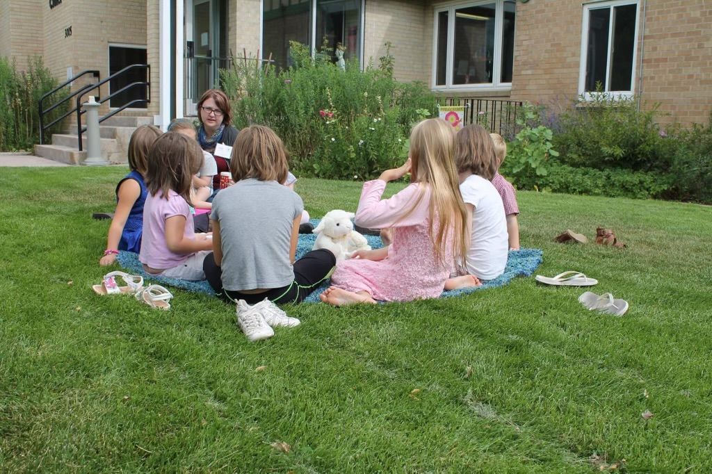 Children sitting in a circle on grass, gathered around a plush toy. Buildings and greenery in the background.