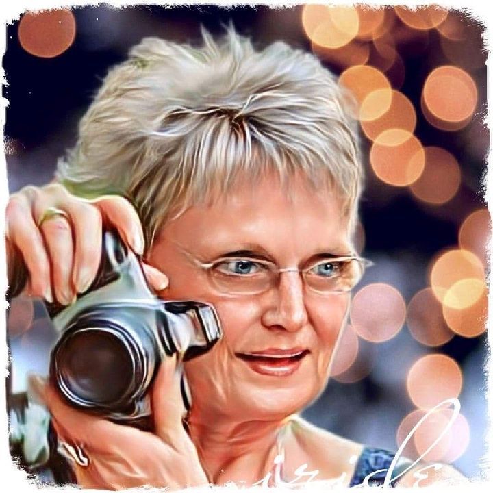 Woman with blonde hair and glasses, holding a camera, smiling, with blurred lights in the background.