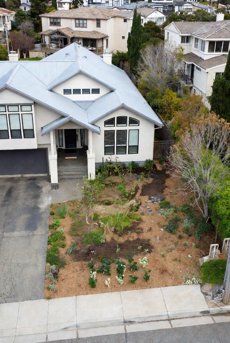 High-angle view of a house with a light-colored exterior and a grey roof, featuring a front garden with mulch and plants.