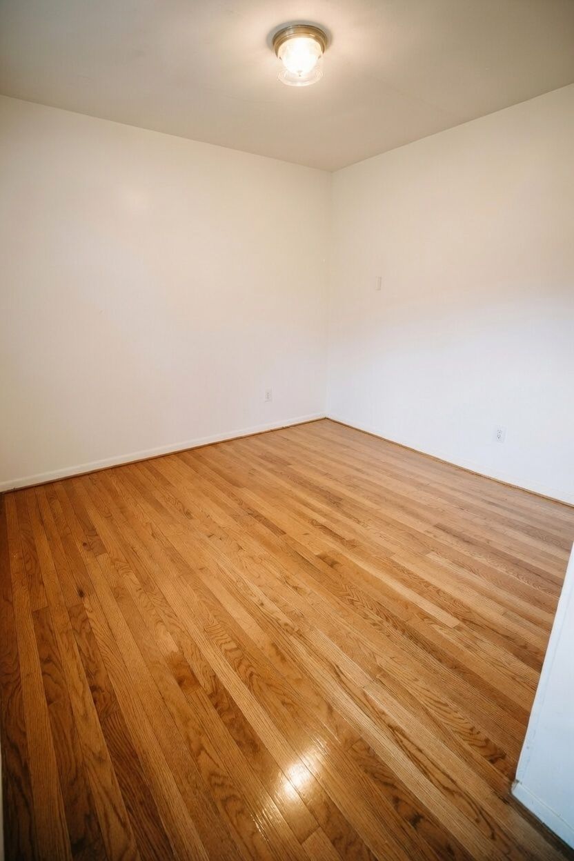 An empty room with white walls and polished light-brown hardwood flooring, illuminated by a ceiling light.