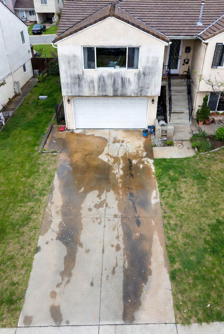 An aerial view of a tan two-story house with a stained driveway and discolored exterior siding.