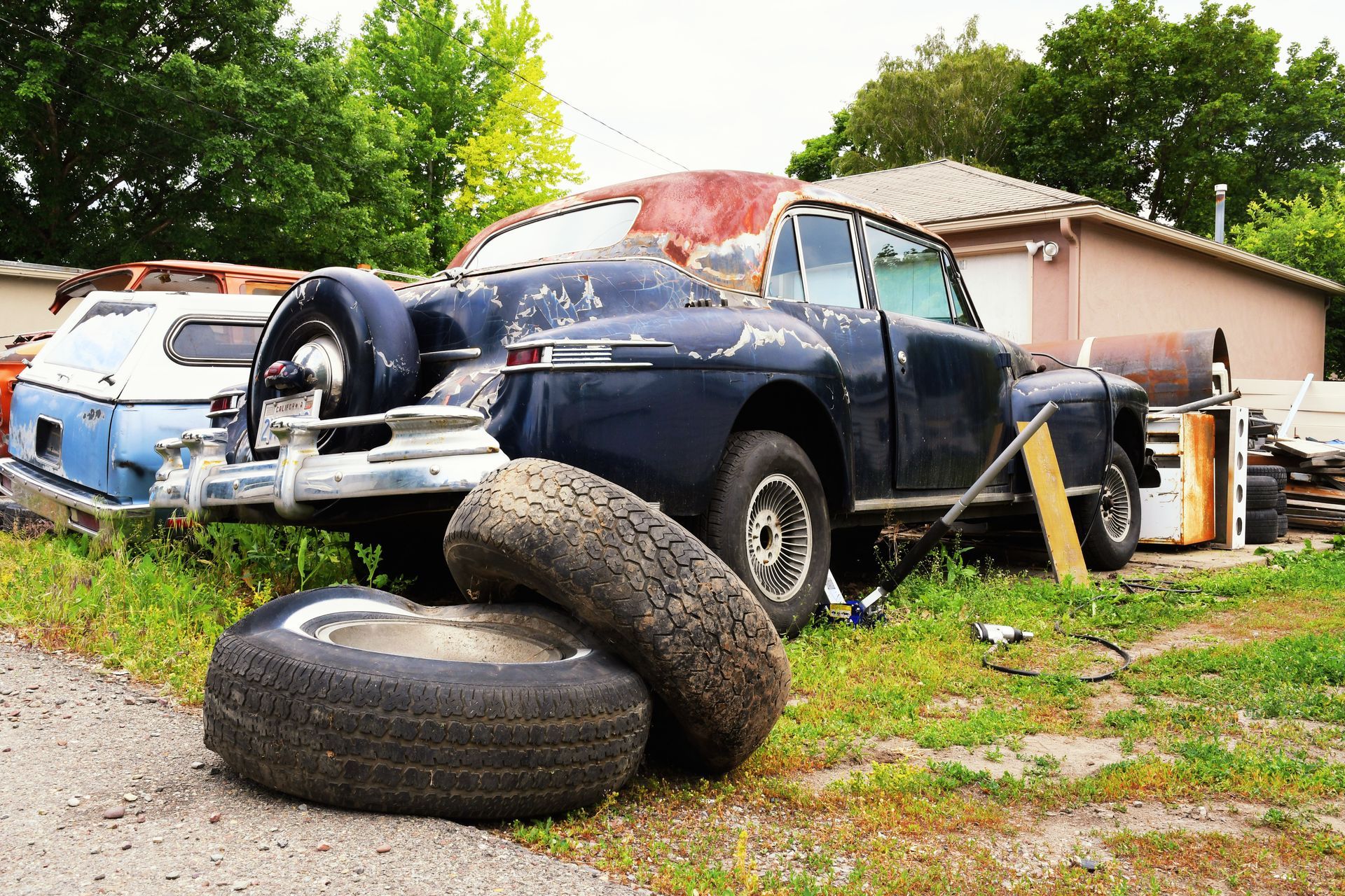 Old, decaying dark blue car in a junkyard, with spare tires in front.