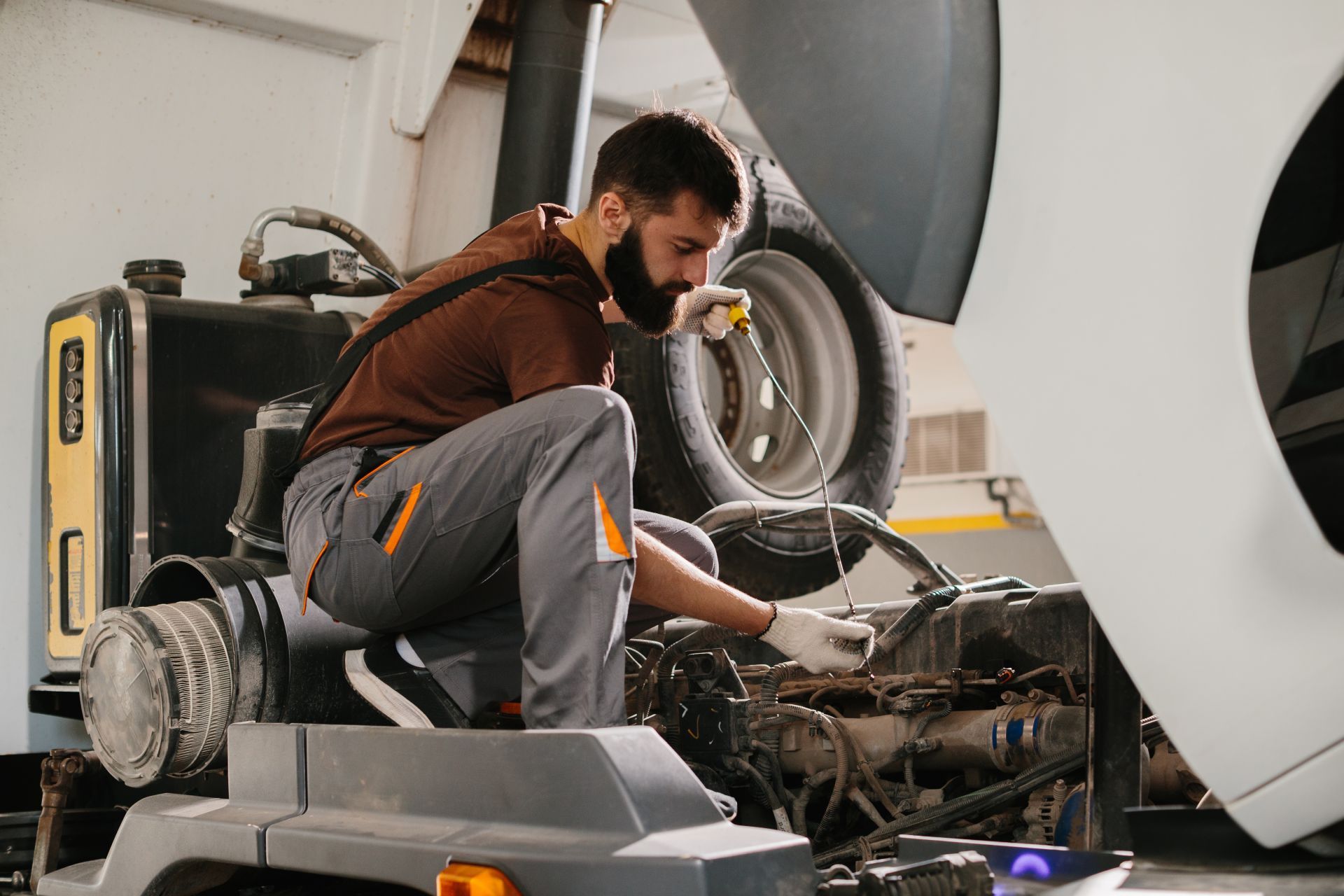 Mechanic examining engine in a garage; wearing gloves, using tool.