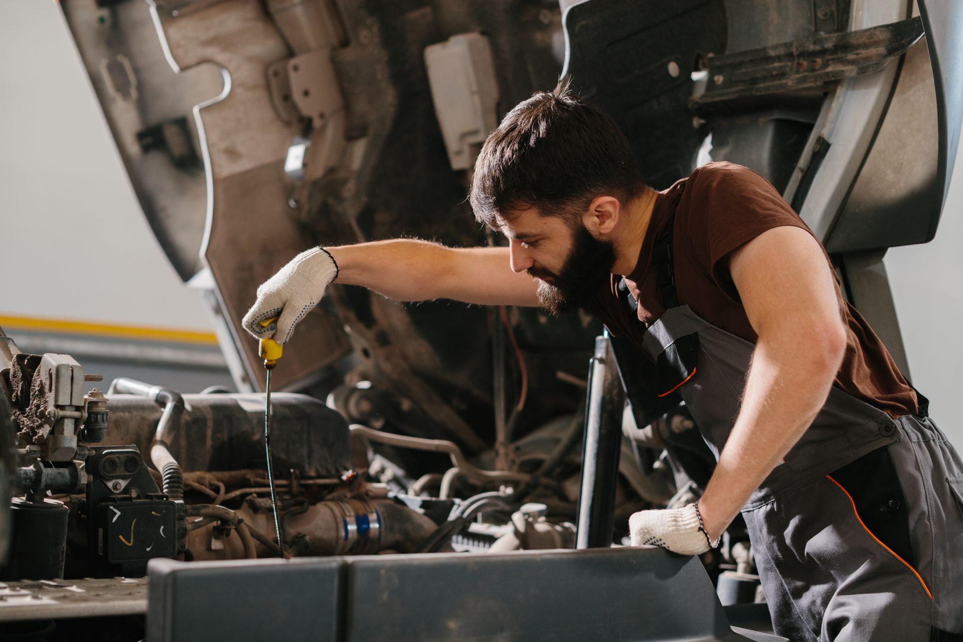 Mechanic in work clothes checking a vehicle's engine with a dipstick in a garage.