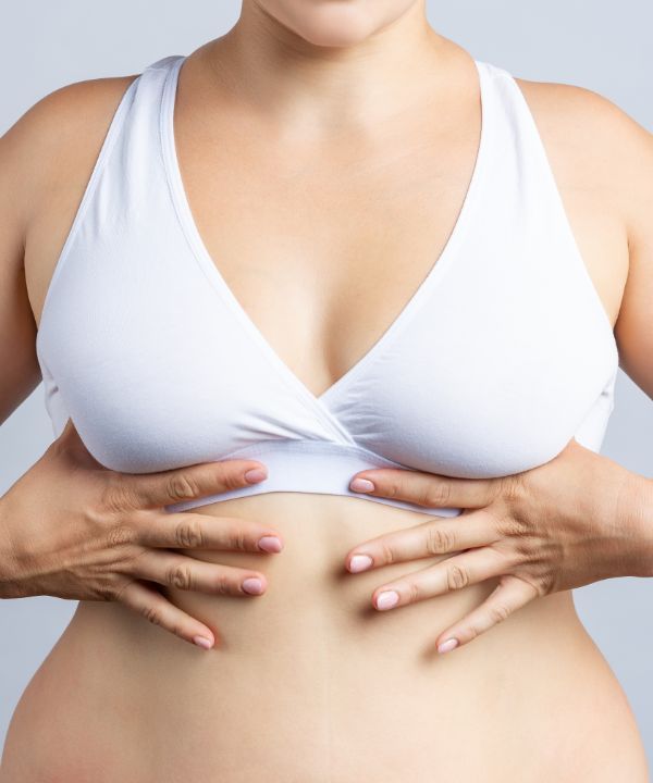 A close-up of a woman wearing a white sports bra, gently holding her abdomen with both hands.