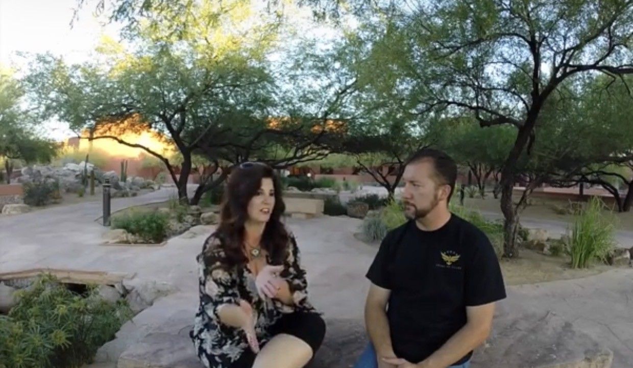 Two people seated outdoors, talking. Woman gestures, wearing floral top. Man in black shirt, listening. Trees and desert landscape in background.