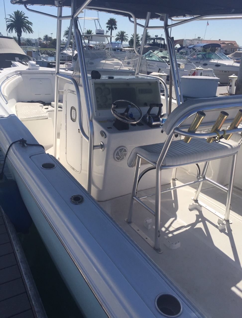 Center console fishing boat docked at a marina on a sunny day, featuring a steering wheel, helm seat, and rod holders.