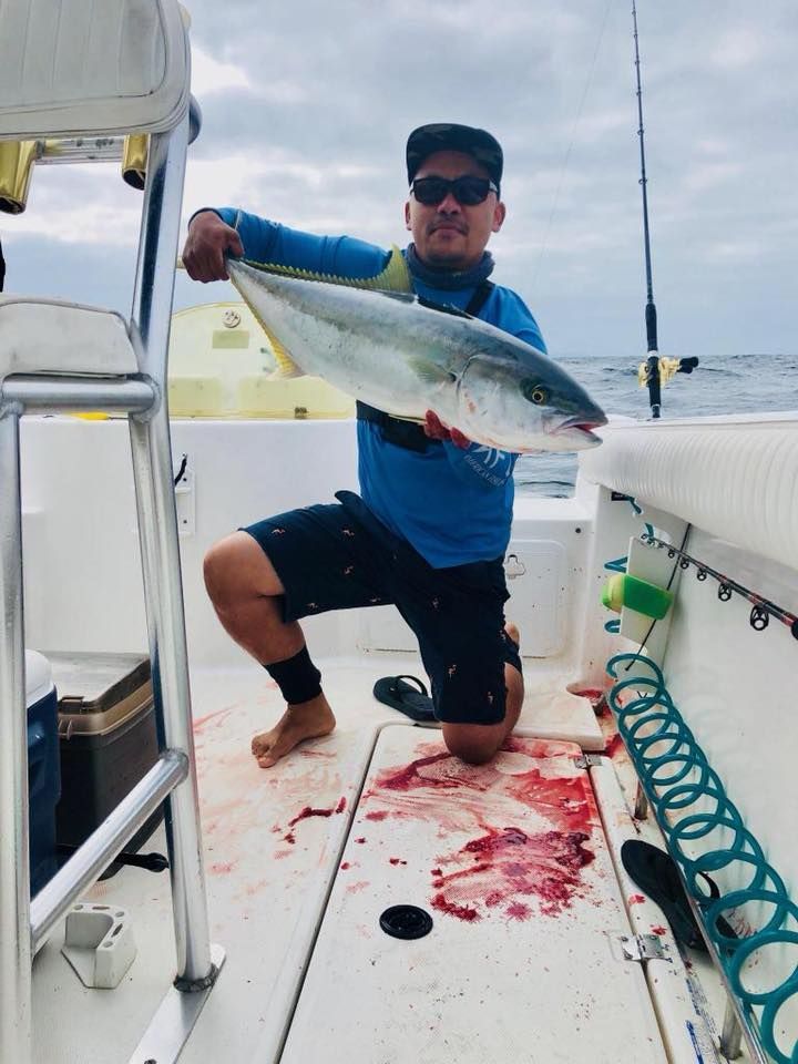 A person kneeling on a boat deck holds a large, silver fish while wearing a blue shirt, sunglasses, and a black hat.