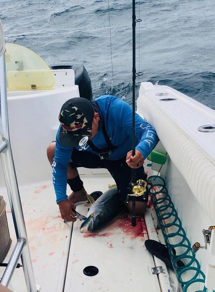 A person in a blue long-sleeved shirt kneels on a boat deck, removing a fishing hook from the mouth of a caught fish.