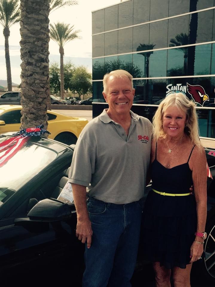 Couple poses next to a black car, in front of a building with palm trees.