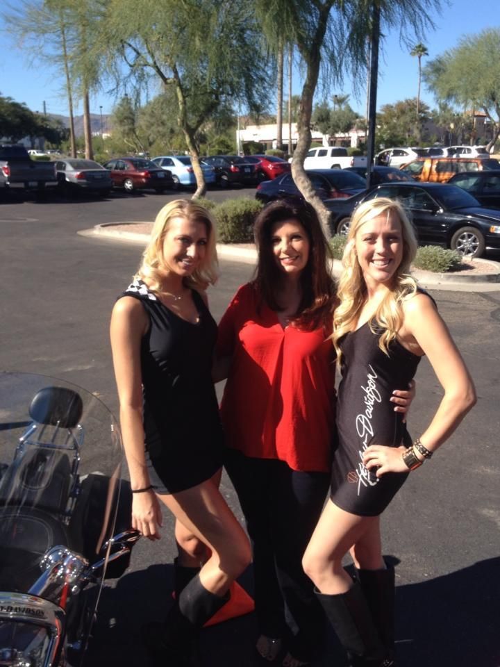 Three people stand together in a parking lot, smiling at the camera, with the woman on the right wearing a branded dress.