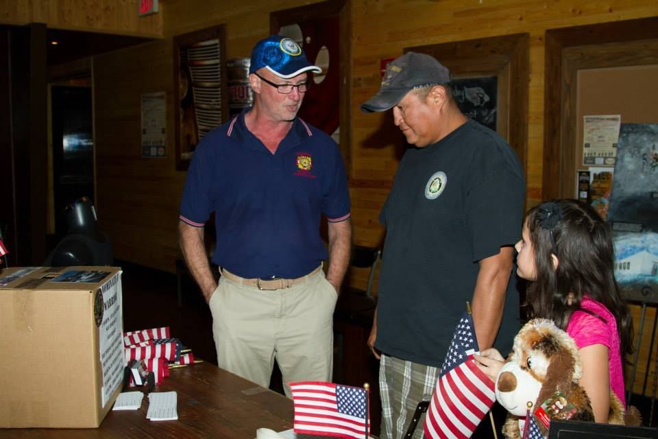 Two people stand with a child holding a stuffed animal near a table displaying US flags in an indoor room with wood walls.